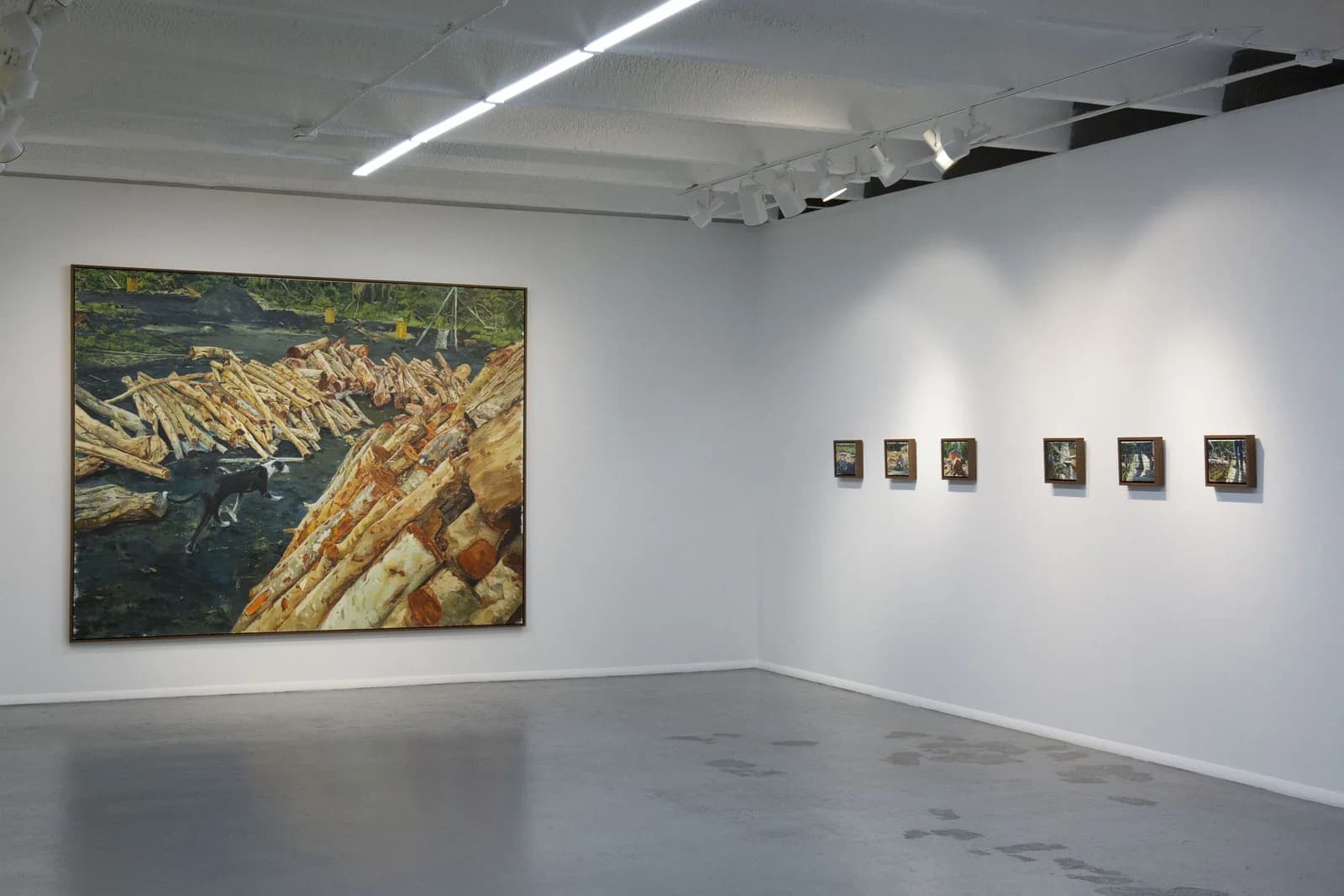 Pintura grande de troncos apilados junto al agua; en la pared contigua, una hilera de seis pinturas pequeñas.