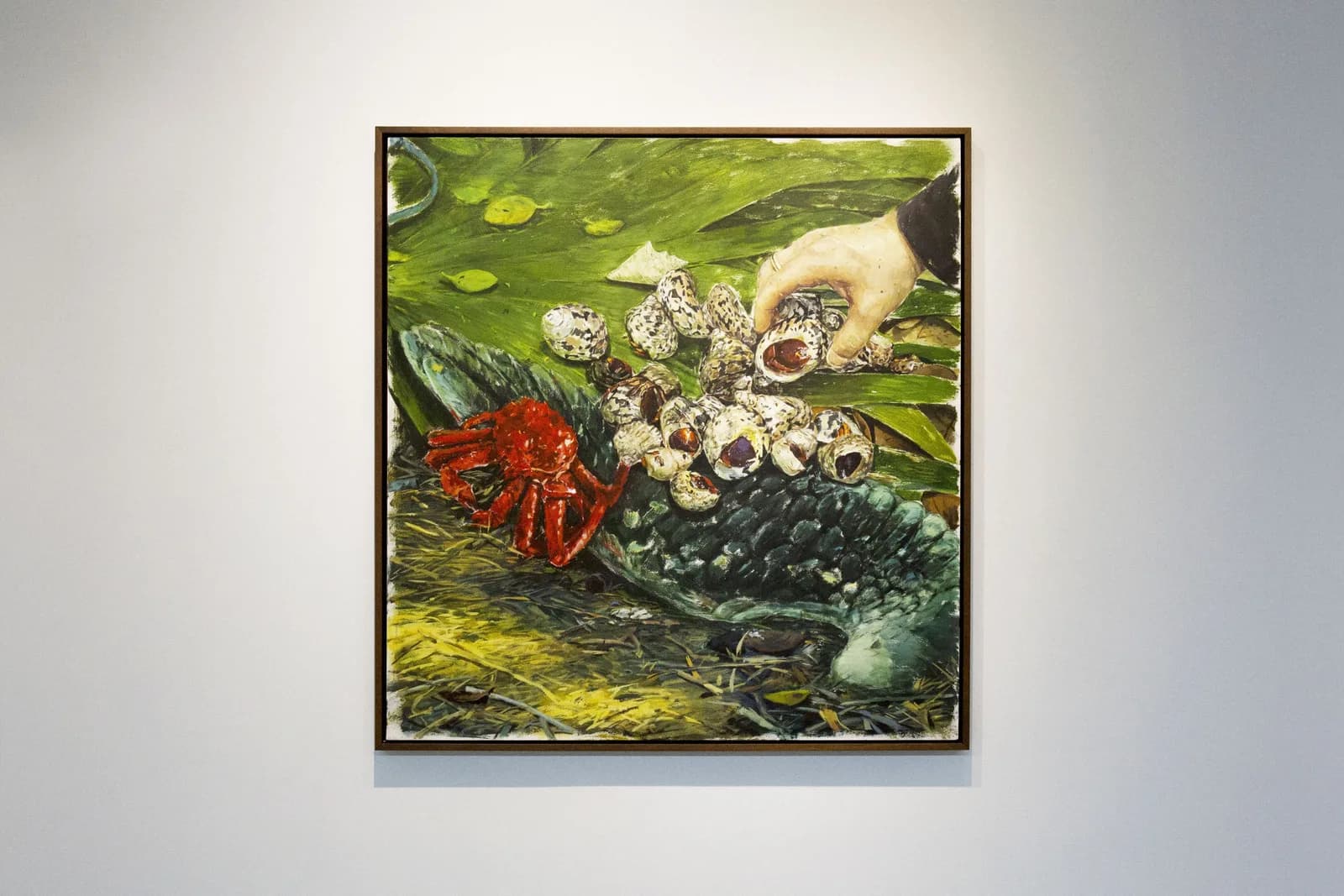Una pintura enmarcada sobre una pared blanca: una mano coloca caracoles junto a un cangrejo rojo, sobre hojas verdes.